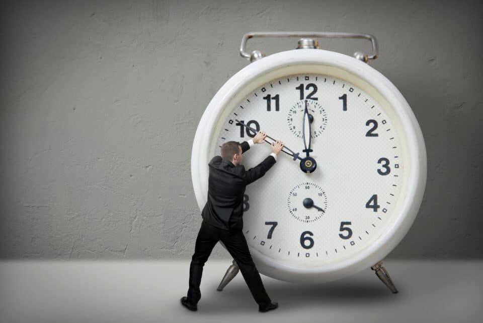 A person in a business suit pushes the hands of a giant clock, symbolising time pressure and the struggle to manage time effectively.