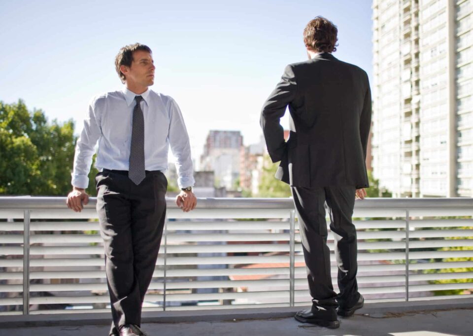 Young adult business man leaning on a balustrade crossing his legs beside his standing colleague while outside an office building.