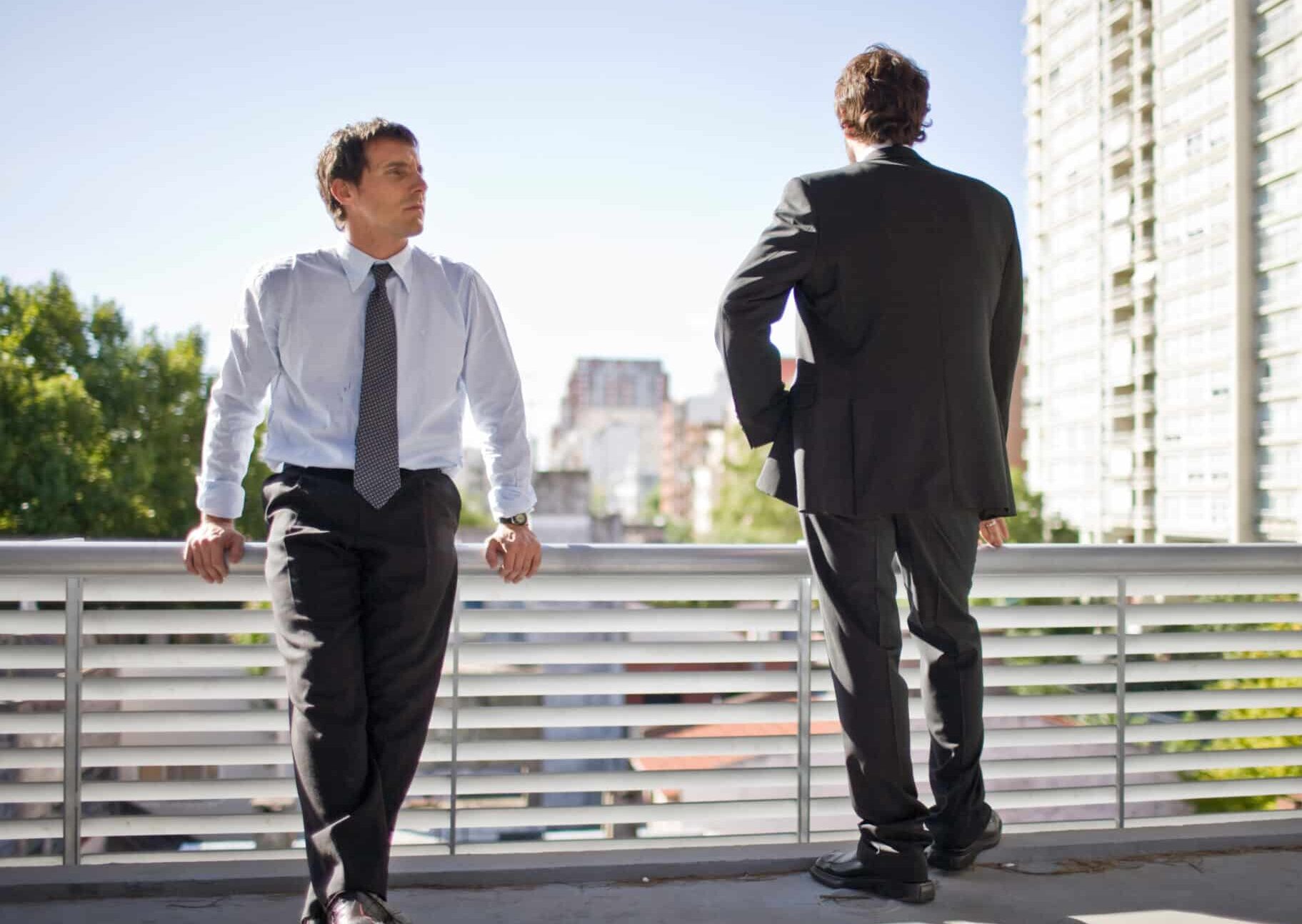Young adult business man leaning on a balustrade crossing his legs beside his standing colleague while outside an office building.