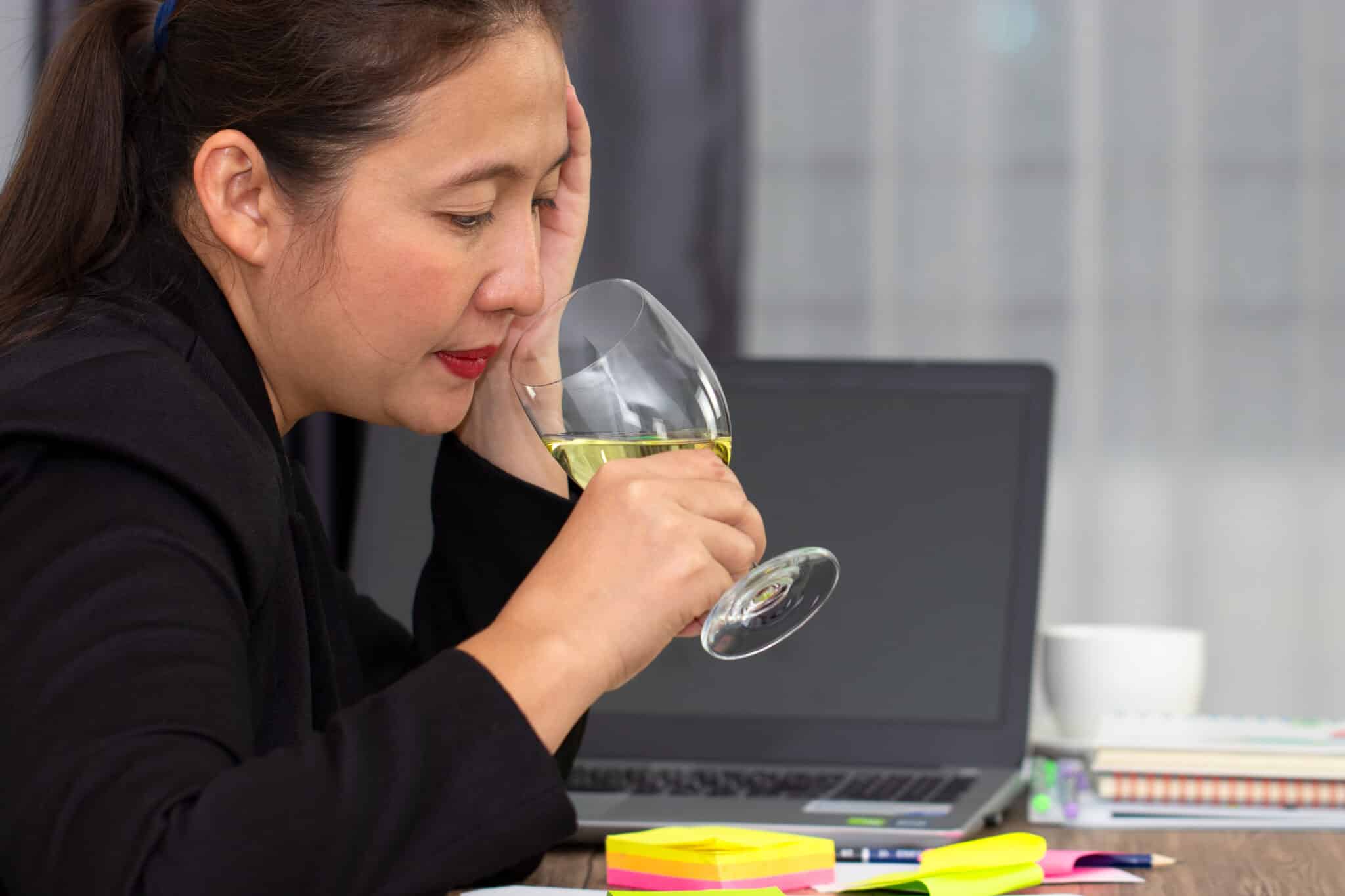 Woman sitting at a desk with a laptop, holding a glass of wine and resting her head in her hand.