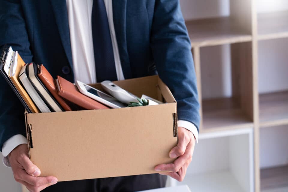 A person in a business suit holding a cardboard box filled with office items, including notebooks, a phone, and folders, suggesting they are leaving or have been laid off.