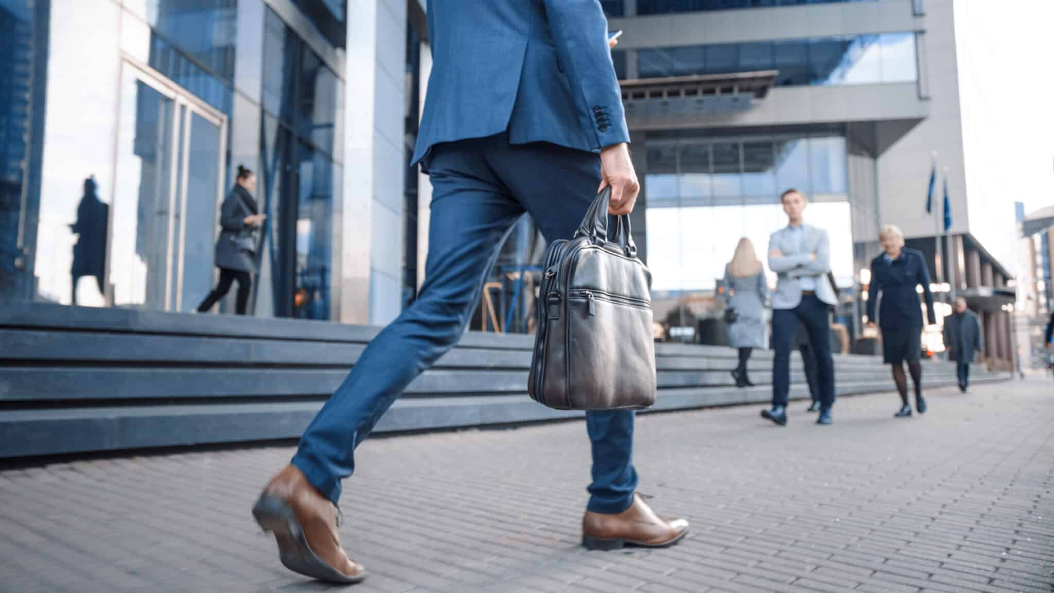 Person wearing a suit and holding briefcase walking in front of a busy office block.