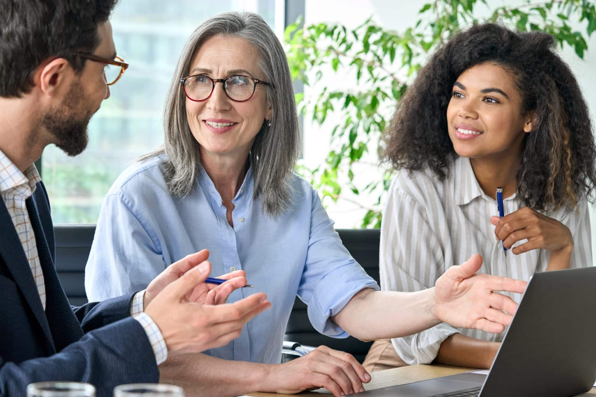 Diverse group of colleagues, including an older woman with gray hair and glasses, engaged in a positive discussion around a laptop in a modern office