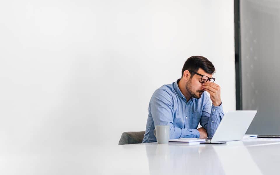 Man in smart office clothes sat at end of desk in from of laptop rubbing eyes with glasses pushed up.