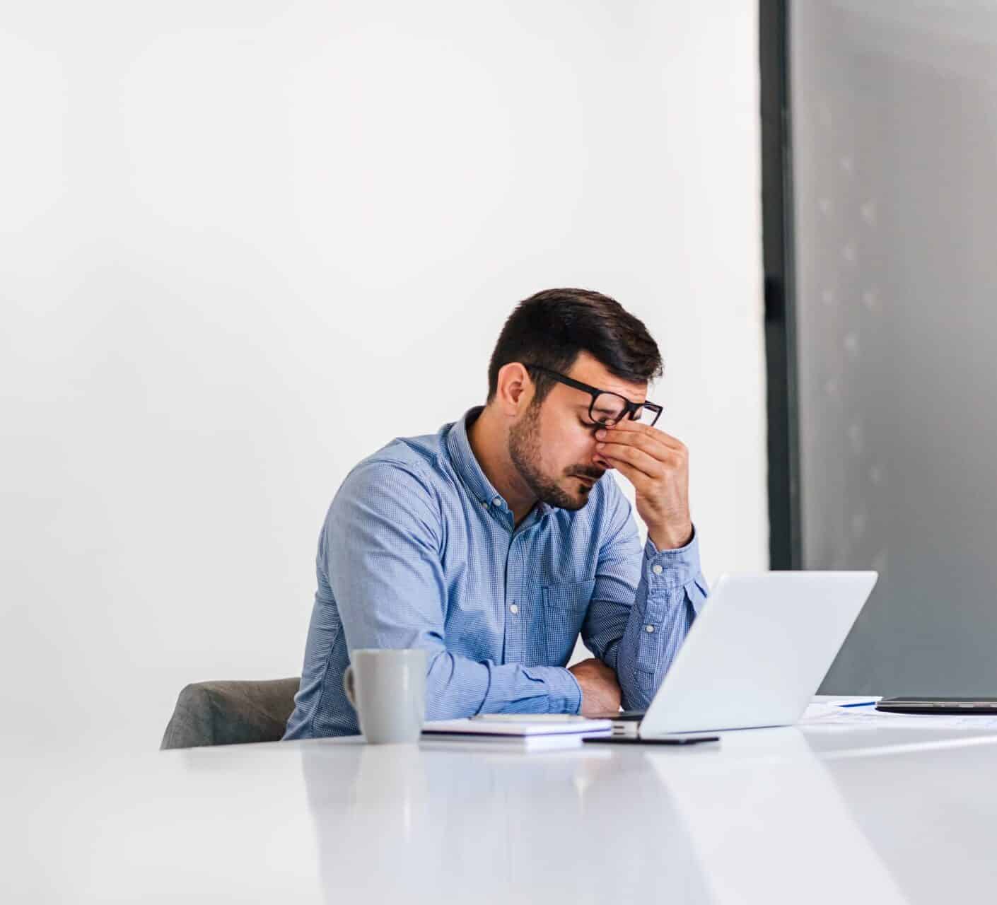 Man sat at desk in office in front of a laptop - looking anxious and tired.