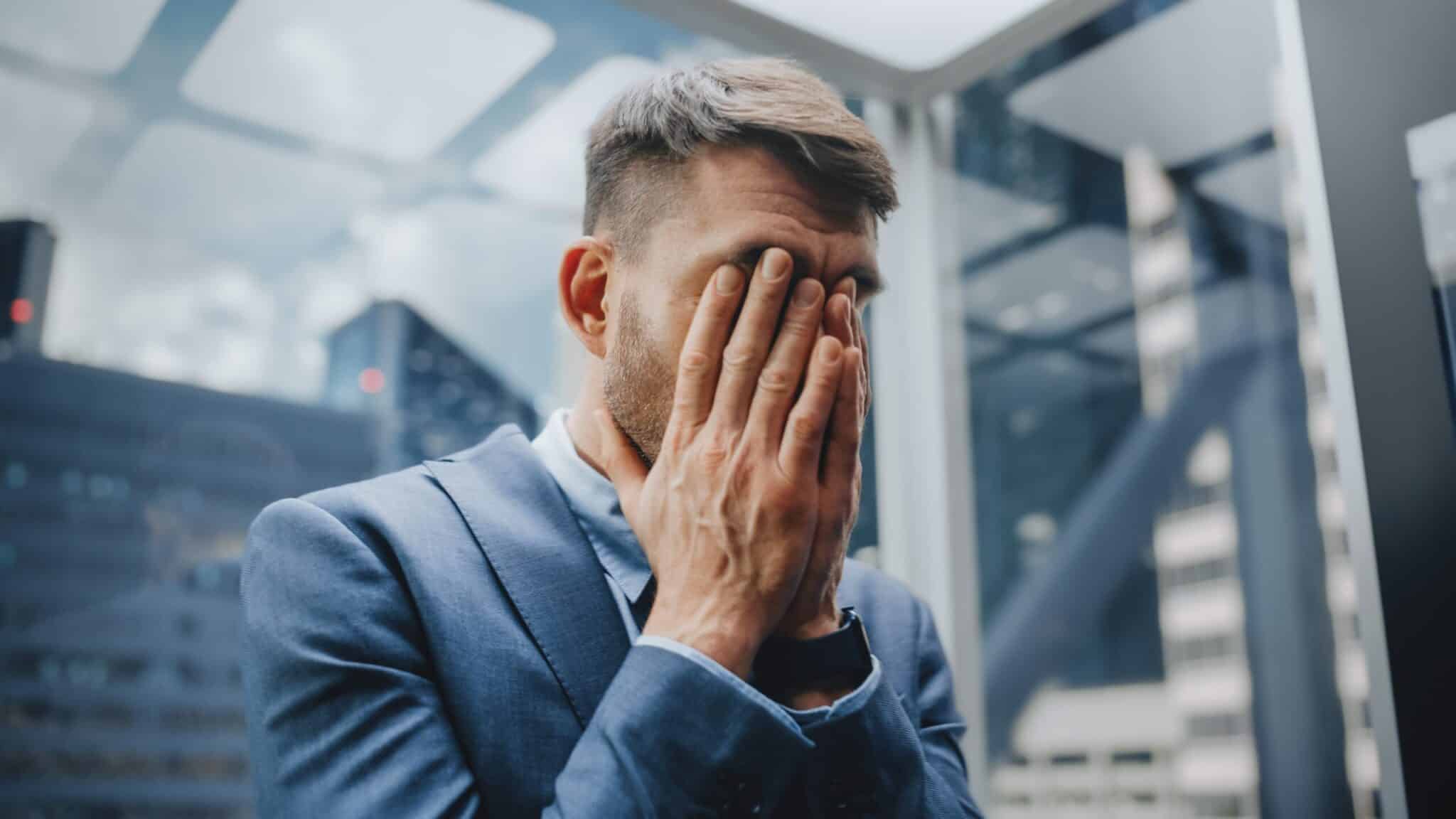 Man in a business suit covering his face with both hands in a glass office, suggesting stress or burnout, with city buildings blurred in the background.