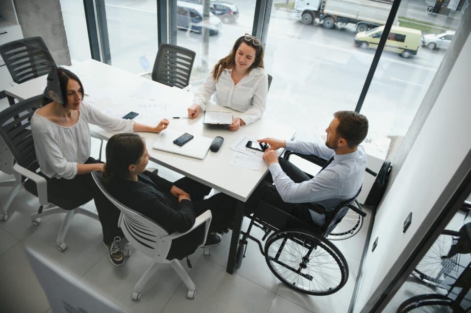 Group of diverse colleagues, including a man in a wheelchair, having a meeting around a table in a modern office with large windows.