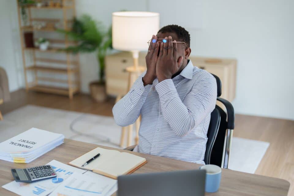 Man sitting at a desk with financial documents, holding his head in his hands in frustration or stress.