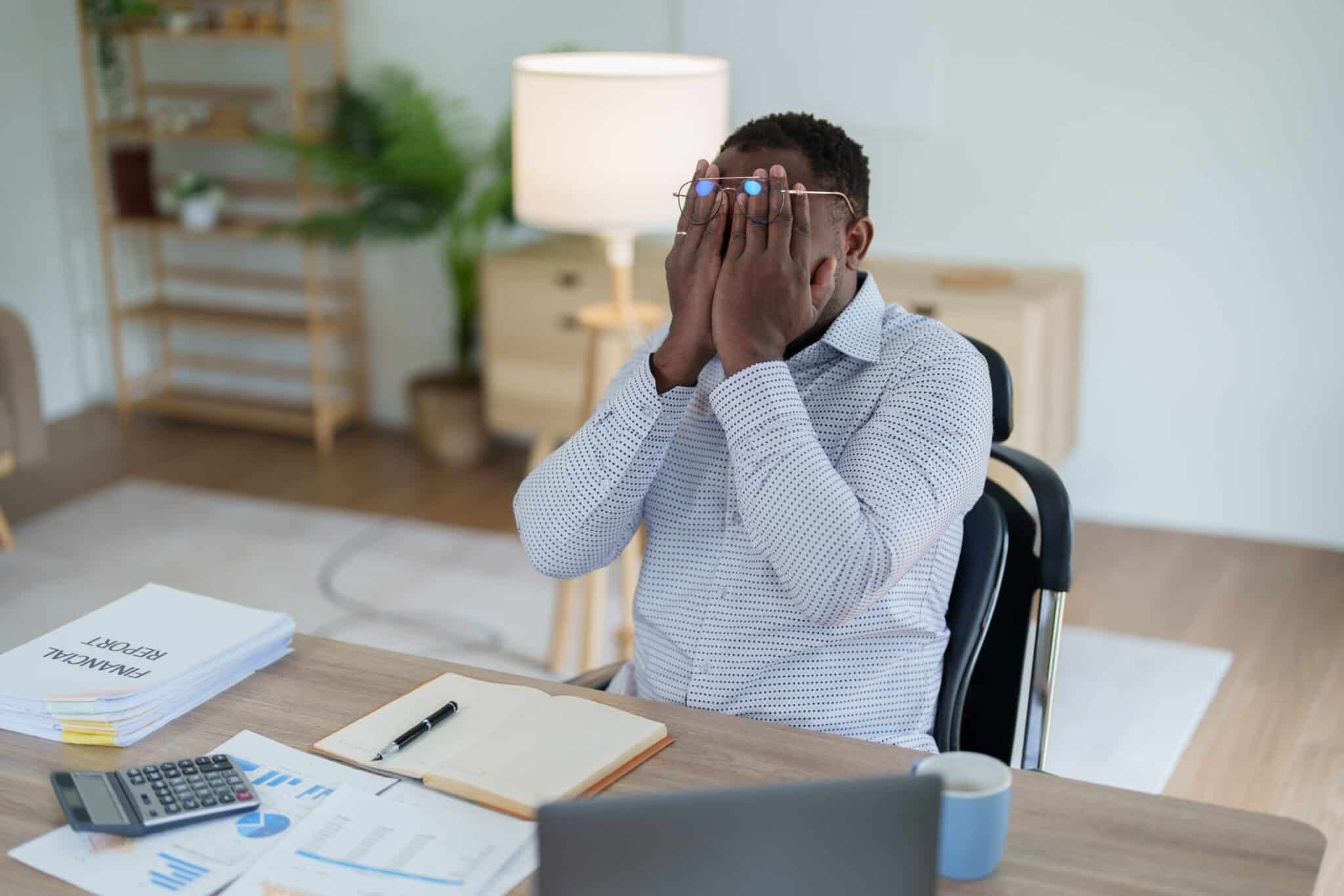 Man sitting at a desk with financial documents, holding his head in his hands in frustration or stress.