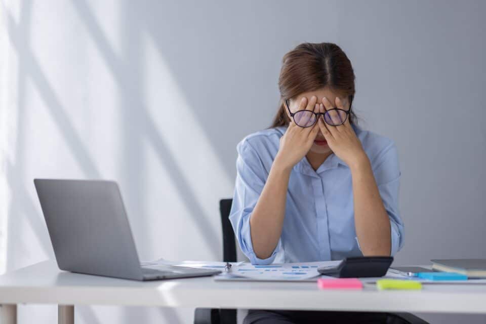 tressed woman sitting at a desk with a laptop and papers, holding her head in her hands.