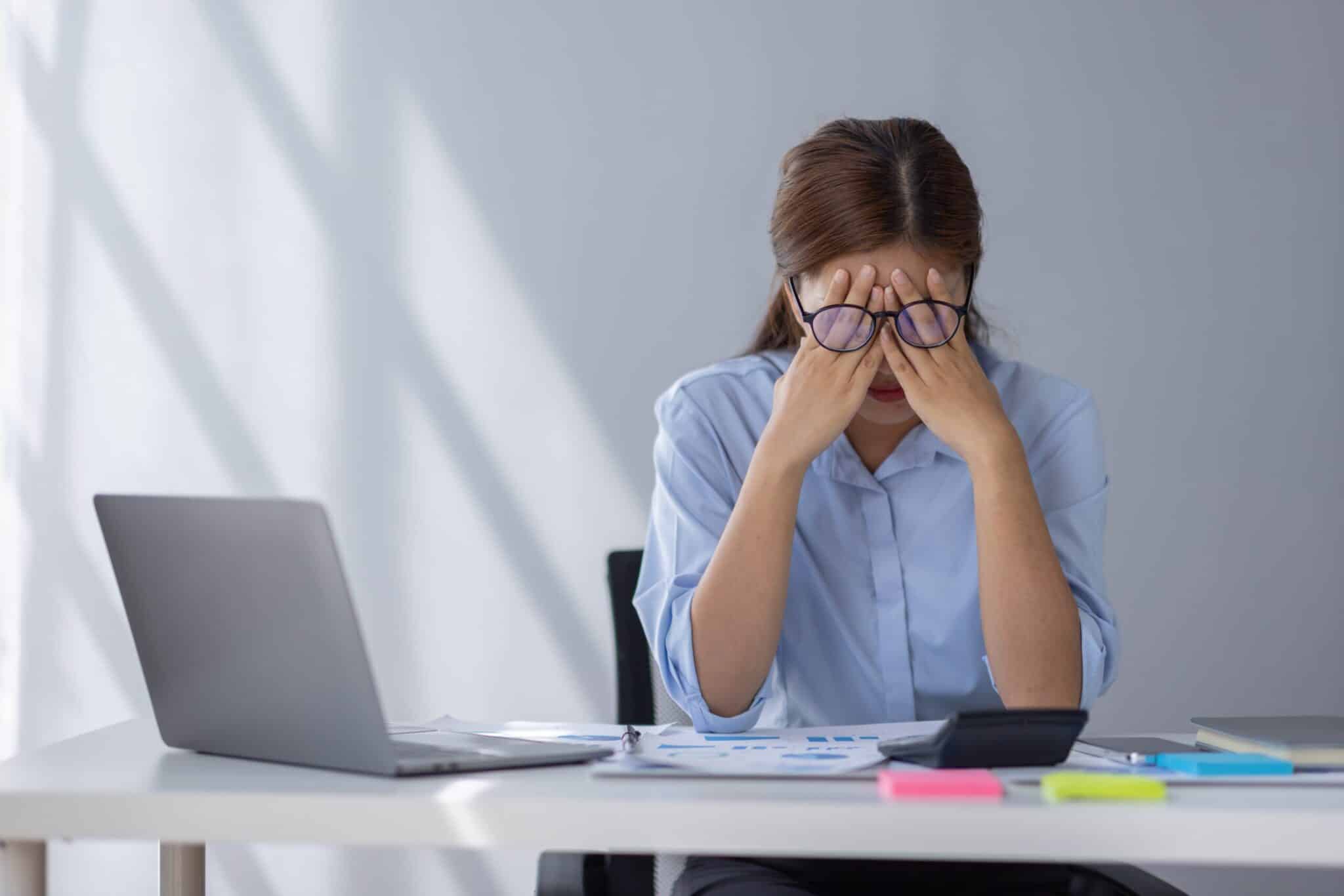 tressed woman sitting at a desk with a laptop and papers, holding her head in her hands.
