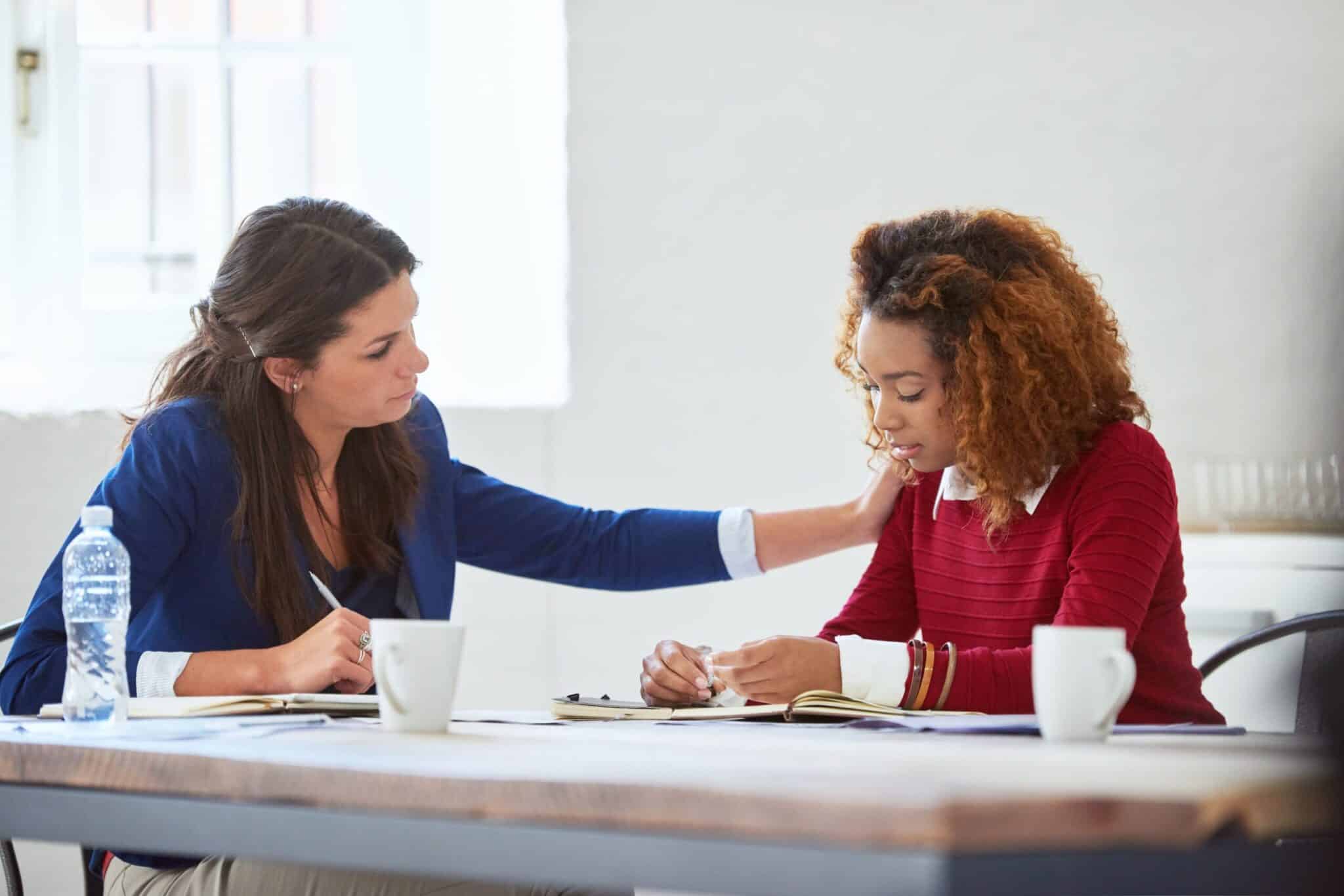 Two women sitting at a table; one woman looks upset while the other gently comforts her with a hand on her shoulder.