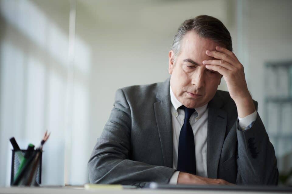 Man in a suit sitting at a desk with his hand on his forehead, looking stressed or worried.
