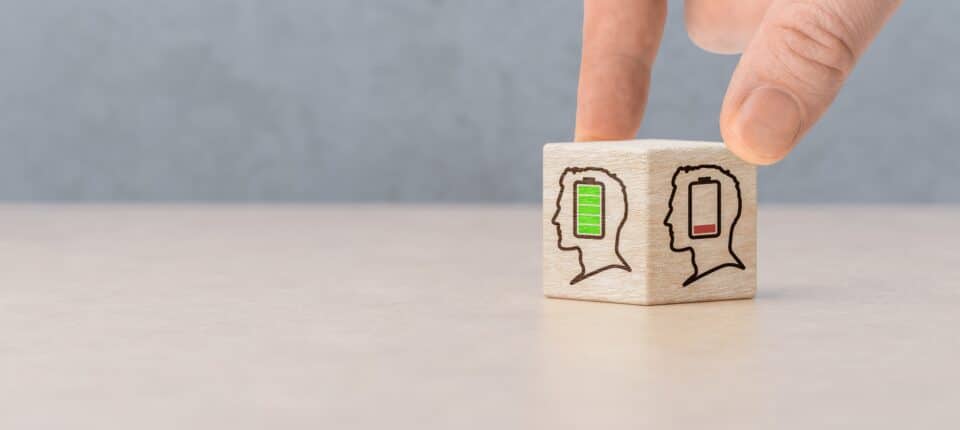 Hand turning a wooden block showing two human profiles, one with a full green battery and one with a low red battery, symbolising mental energy and wellbeing.