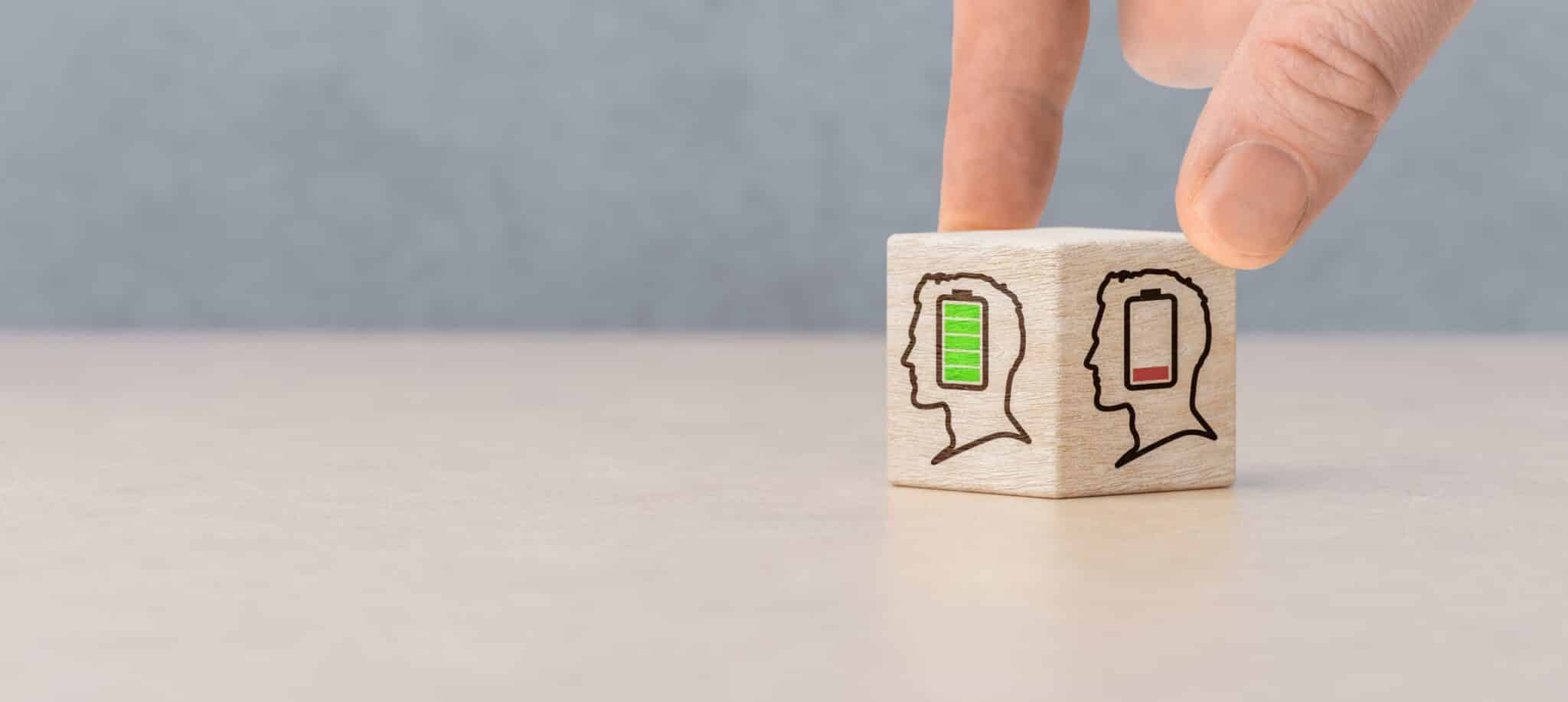 Hand turning a wooden block showing two human profiles, one with a full green battery and one with a low red battery, symbolising mental energy and wellbeing.