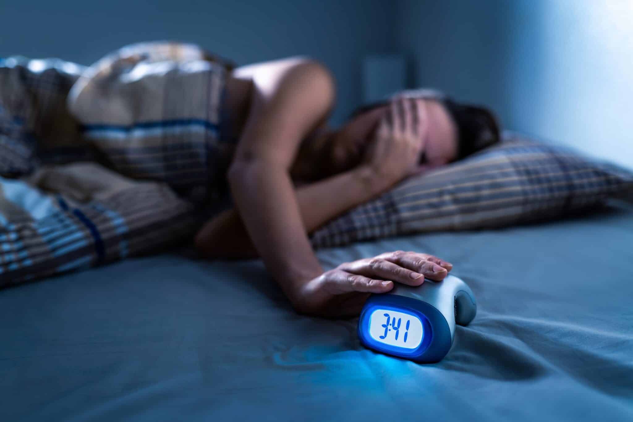 Person lying awake in bed at night, covering their face with one hand while the other rests on an alarm clock showing 3:41 a.m