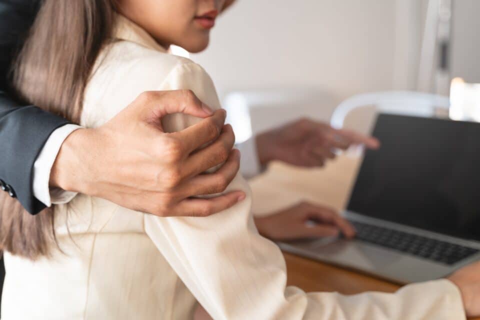 Person placing a hand on a colleague’s shoulder while they work at a laptop, suggesting unwanted physical contact in a workplace setting.