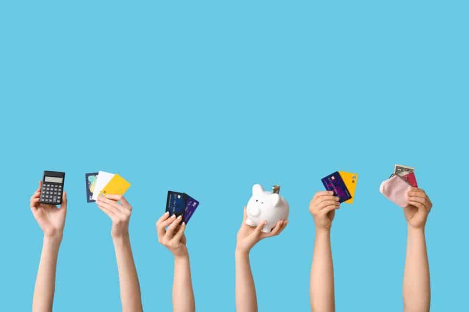 Multiple hands holding up a calculator, credit cards, a piggy bank, and a coin purse against a blue background, symbolizing personal finance and money management.