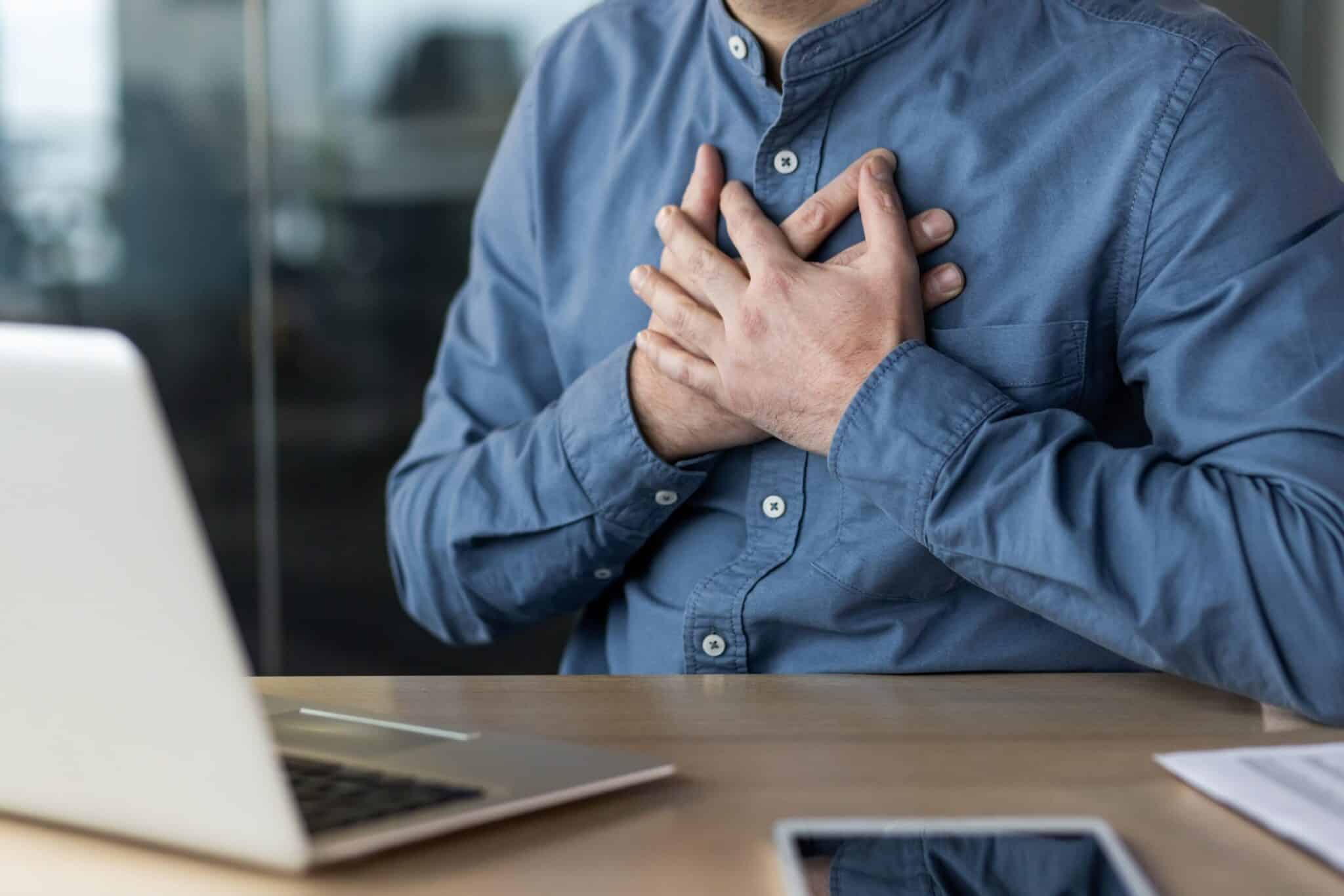A man in a blue shirt sits at a desk with a laptop, holding both hands over his chest, suggesting he may be experiencing chest pain or emotional stress.