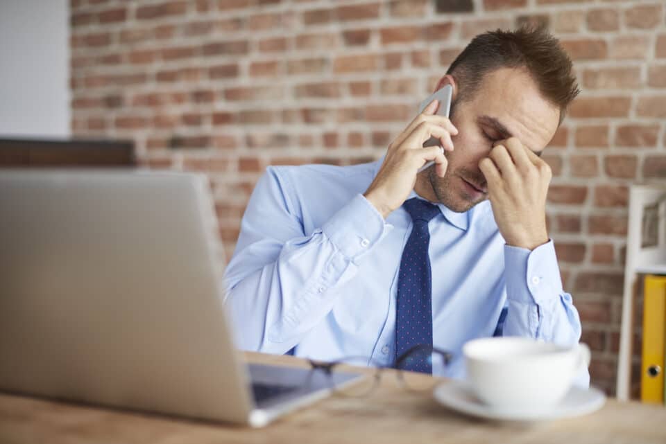 A man in a blue shirt and tie sitting at a desk with a laptop and coffee cup, holding a phone to his ear while pinching the bridge of his nose, looking stressed or fatigued.