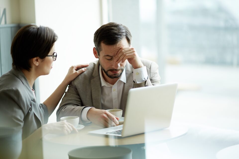 Woman comforting a stressed man sitting at a table with a laptop and coffee, as he holds his head in his hand.