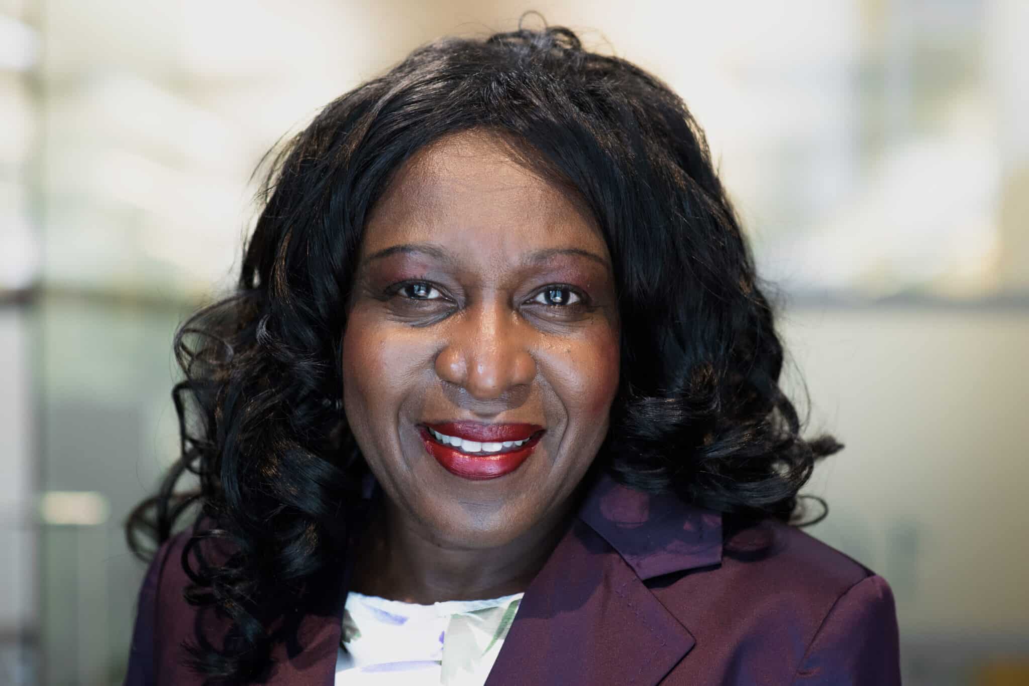 Smiling woman in a purple blazer with dark curly hair, wearing red lipstick, photographed in a professional indoor setting.