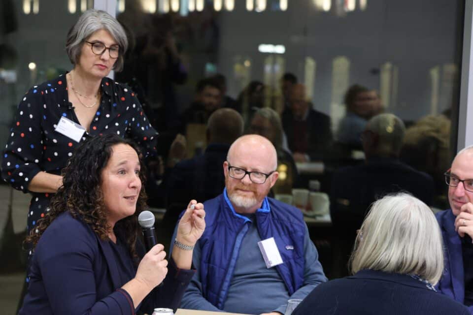 Woman speaking into a microphone during a discussion at an event, with others seated around a table and one woman standing nearby.