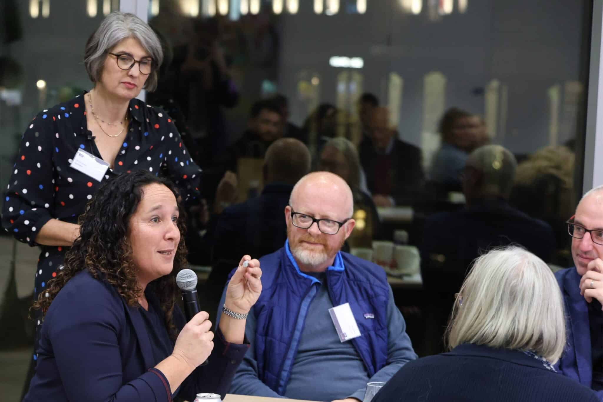 Woman speaking into a microphone during a discussion at an event, with others seated around a table and one woman standing nearby.