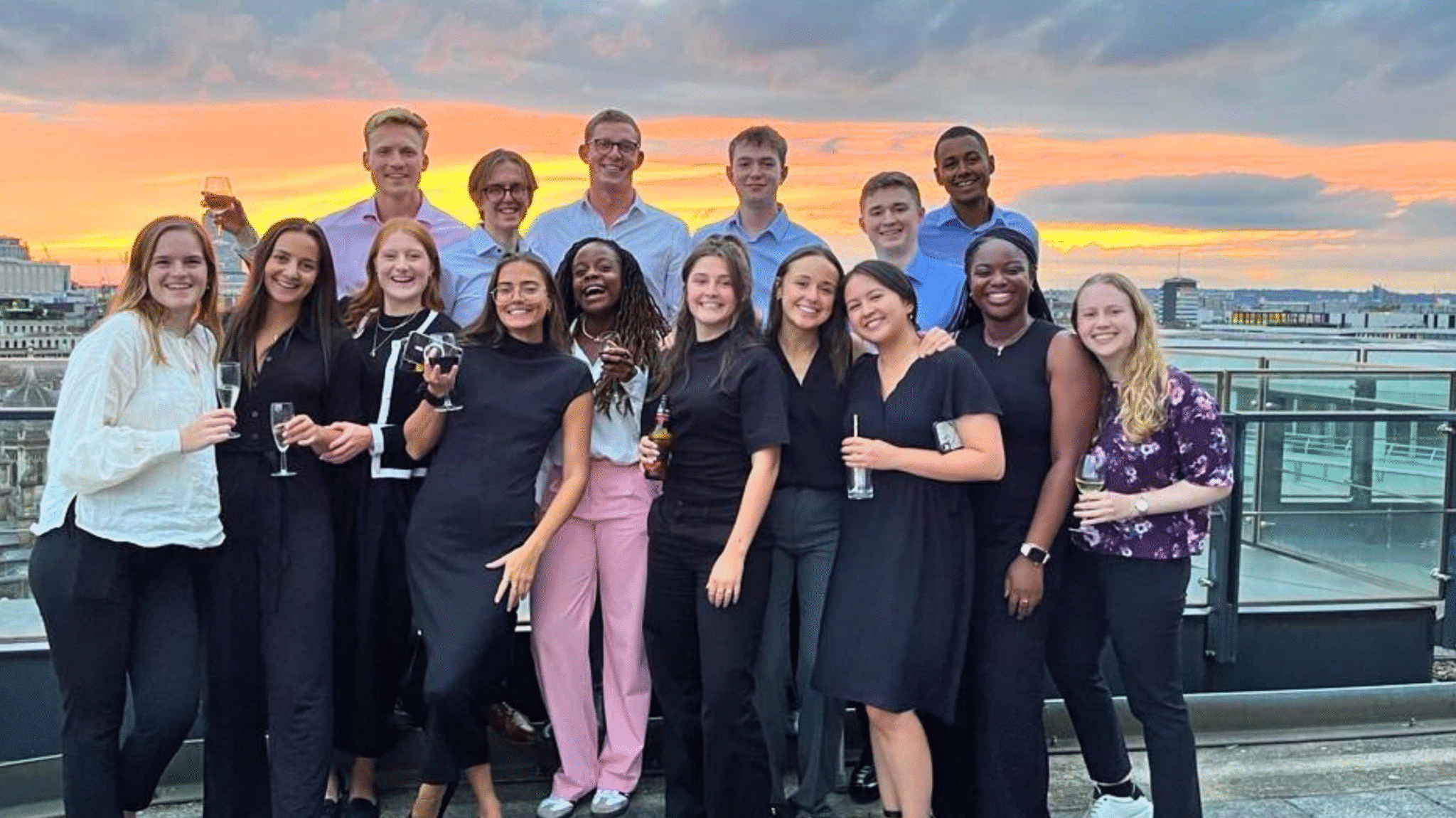 A group of smiling young professionals stand together on a rooftop at sunset, holding drinks and posing for a photo, with a city skyline and dramatic sky in the background.