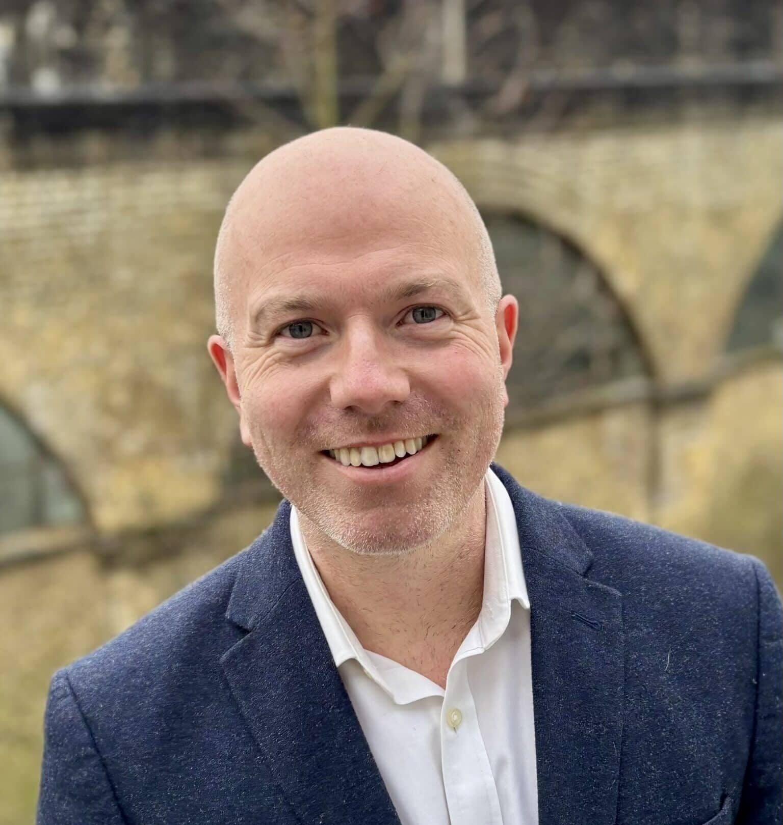 A smiling person with a shaved head wearing a navy blazer and white shirt, standing outdoors in front of an old stone wall with arched windows.