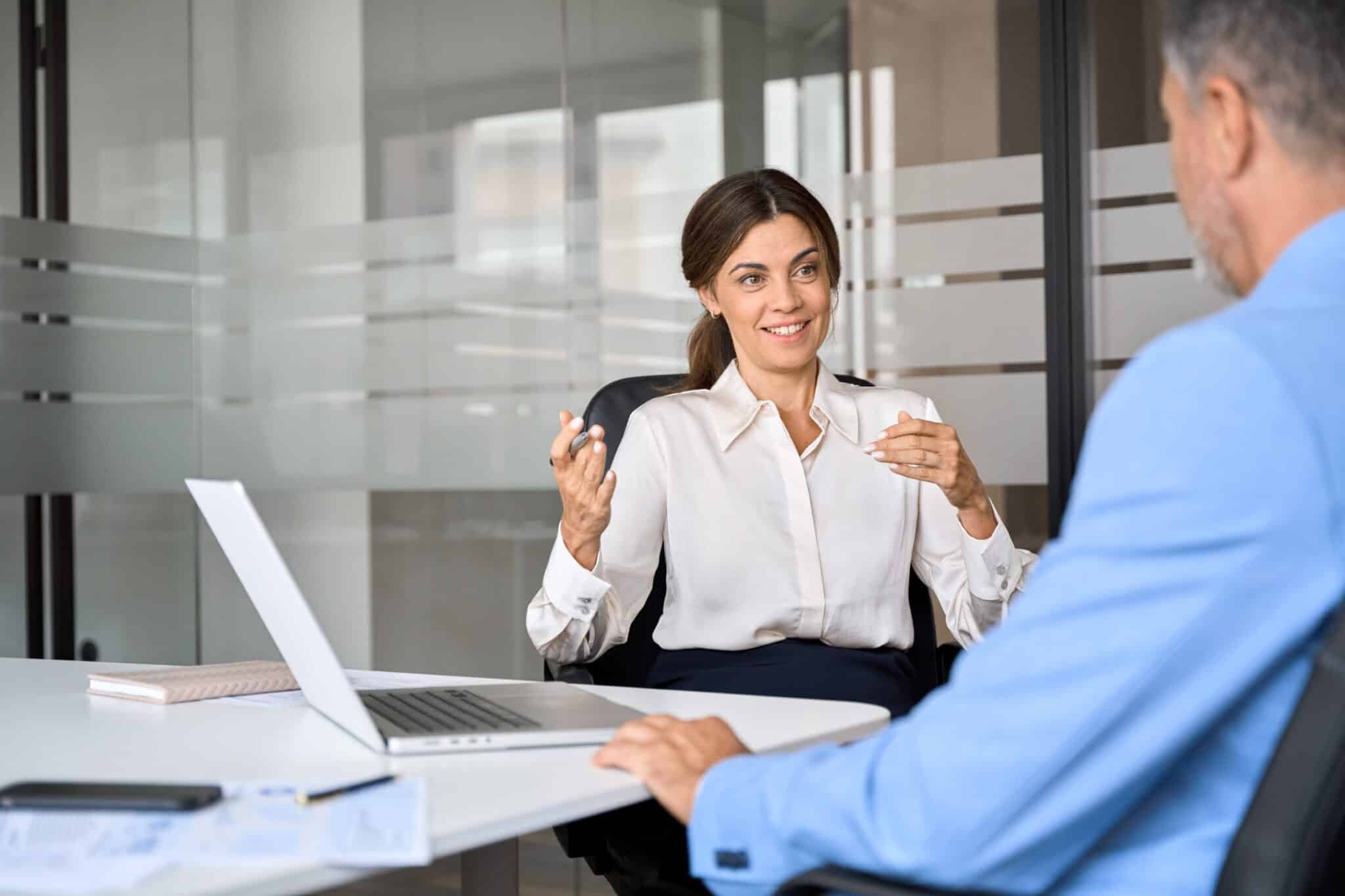 two professionals sat at a desk with a laptop on it. They are talking.