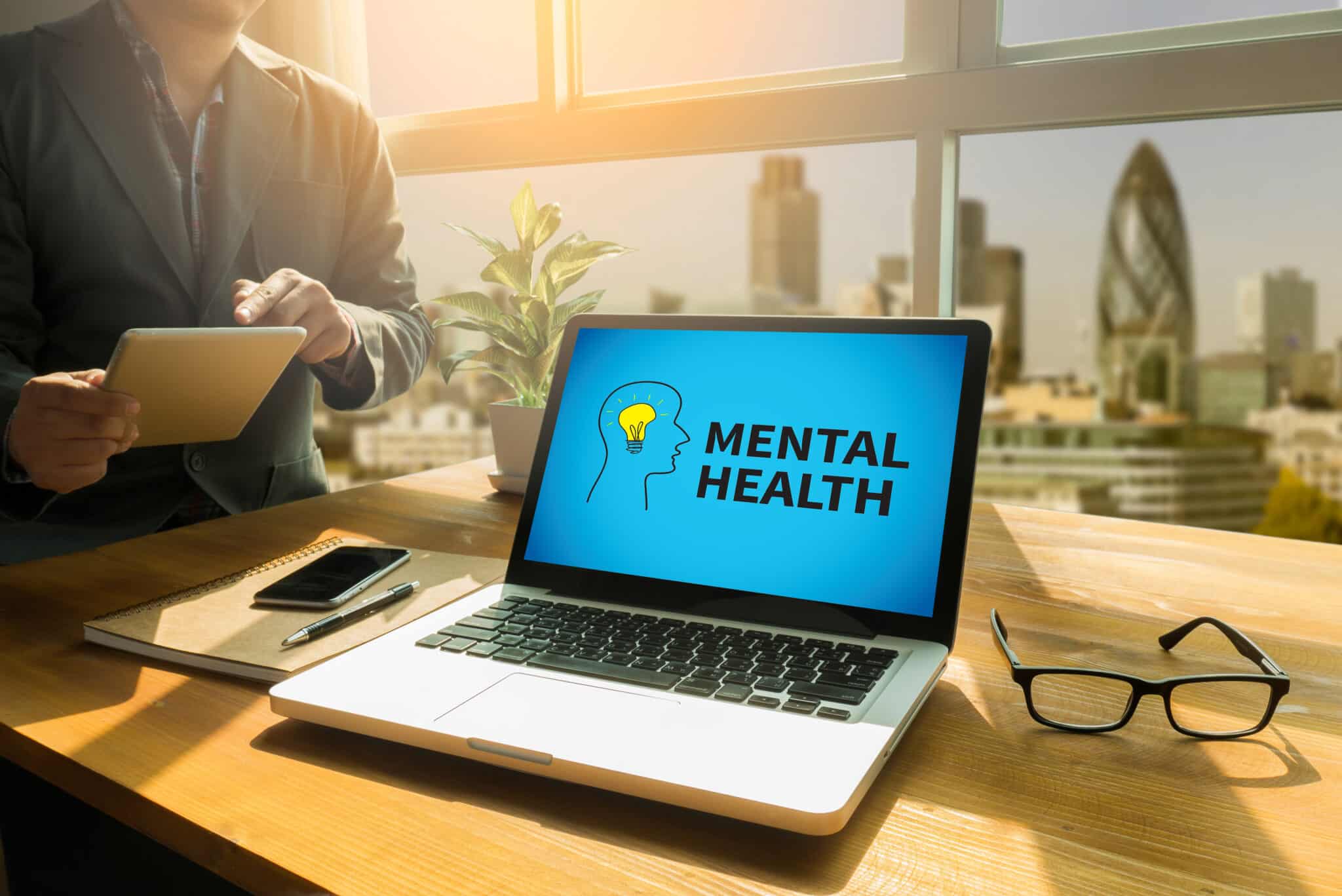 Laptop screen displaying the words “Mental Health” on a desk with a tablet, phone, and glasses.