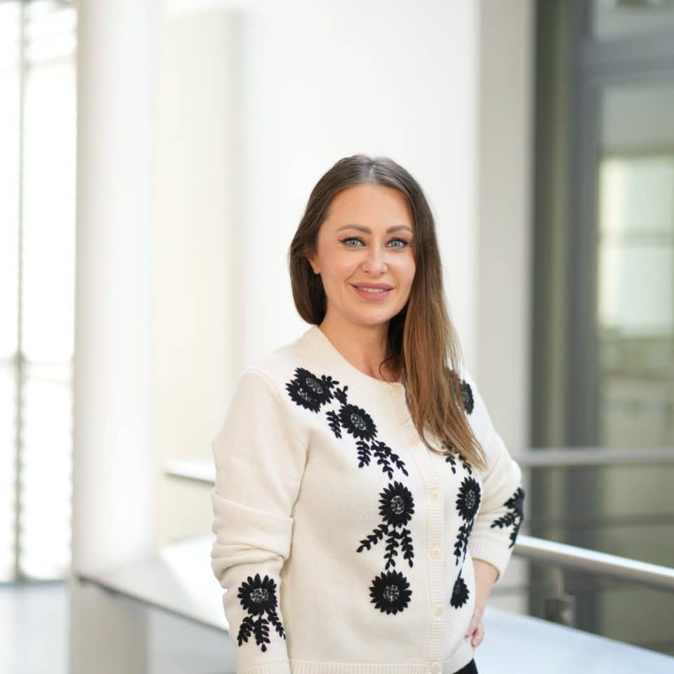 A woman standing indoors, smiling at the camera. She has long brown hair and is wearing a cream-coloured cardigan with black floral embroidery. The background is softly lit with large windows and modern architectural details.