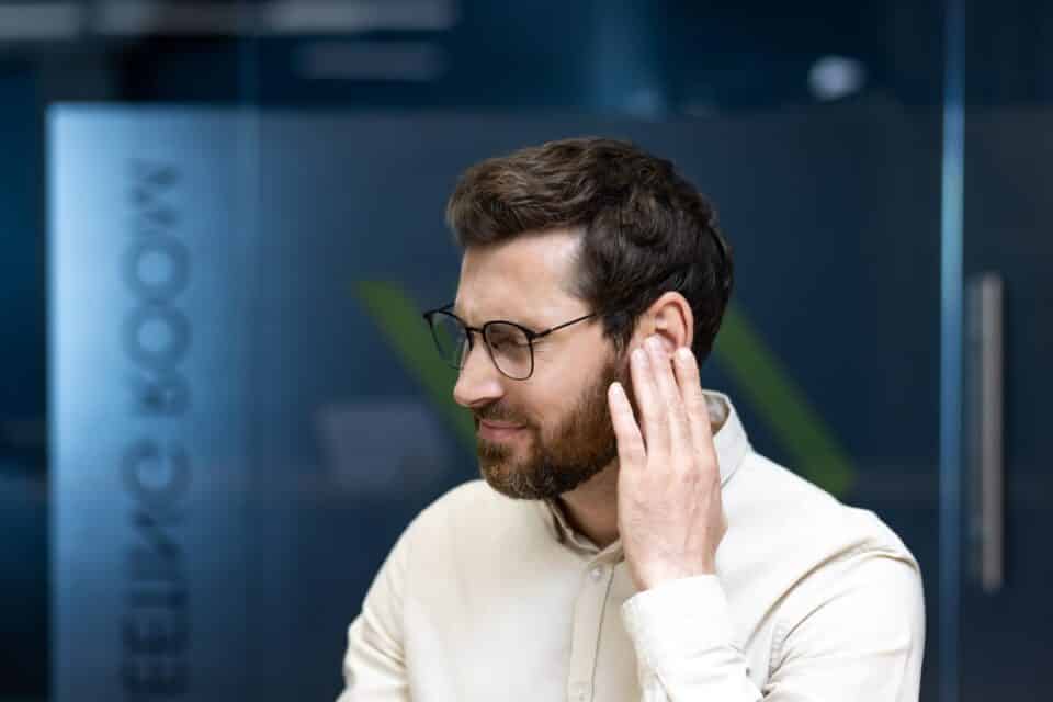 A man wearing glasses touches his ear with a concerned expression while sitting in an office setting