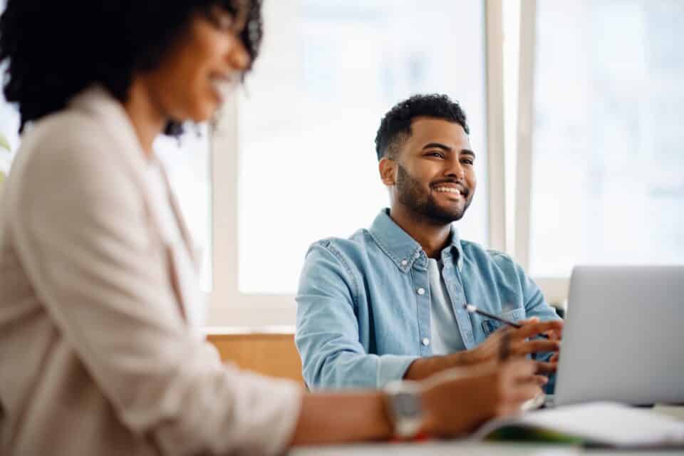 A smiling man seated at a desk using a laptop in a modern office, with another person working nearby.