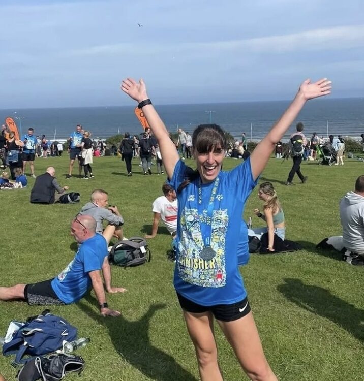 Woman wearing a blue finisher’s running shirt and medal stands on grass with arms raised in celebration after a race, with other runners and the sea visible in the background.
