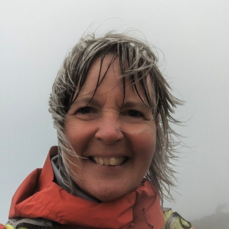Smiling woman outdoors wearing a waterproof jacket, with windblown hair and a cloudy sky in the background.