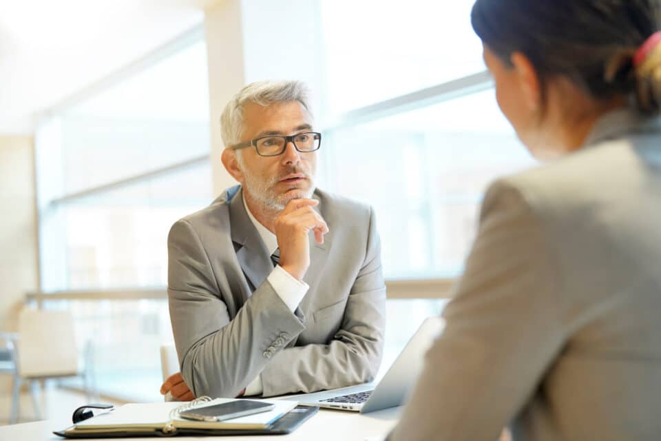 Two professionals in conversation at a desk, with one person listening attentively during a workplace discussion.