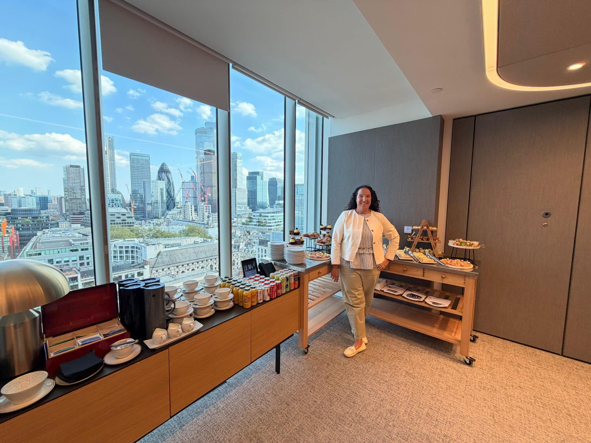 Person standing beside a refreshment table in a modern office meeting room, with large windows overlooking the London skyline, including the Gherkin.