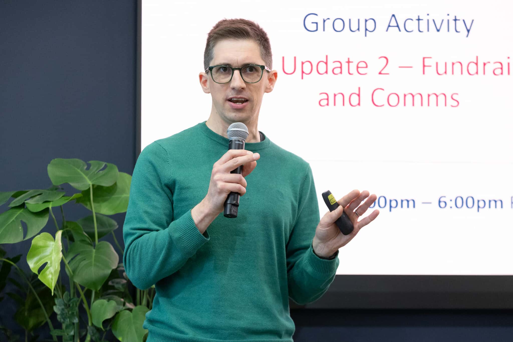 Man in green jumper giving a talk in front of an audience with a presentation in the background