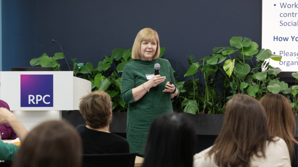 Trish McLellan from LawCare speaks to an audience at an event, holding a microphone and presentation remote. She stands beside a lectern with the RPC logo, with large green plants behind her, while attendees listen from their seats.