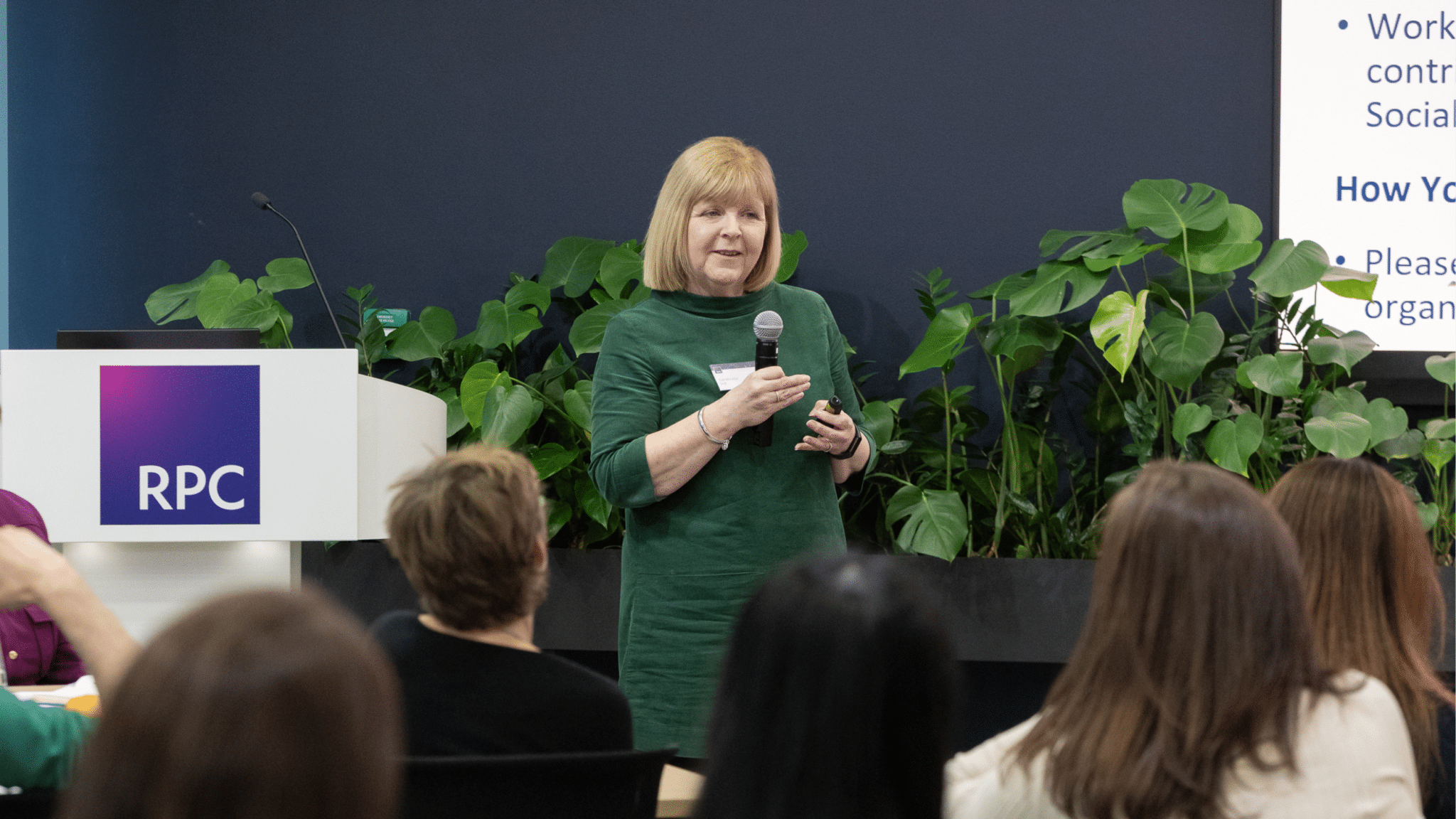 Trish McLellan from LawCare speaks to an audience at an event, holding a microphone and presentation remote. She stands beside a lectern with the RPC logo, with large green plants behind her, while attendees listen from their seats.