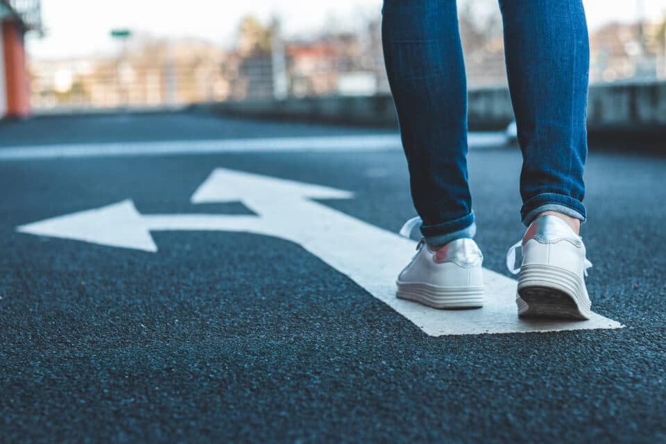 Person standing on a road beside a white directional arrow that splits into two paths, symbolising making a choice or deciding between different directions.
