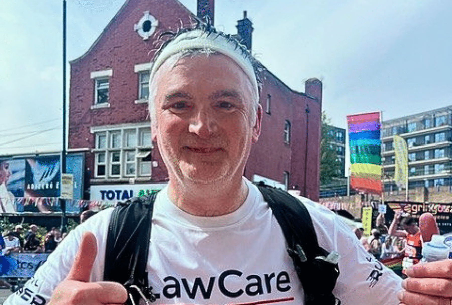 A smiling participant wearing a LawCare T-shirt and backpack gives two thumbs up while standing in a busy street during a Pride event. Rainbow flags and spectators line the route, with buildings and colourful banners visible in the background.