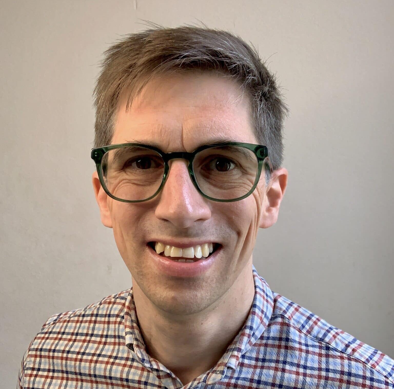 Head-and-shoulders portrait of a smiling man wearing green-framed glasses and a checked shirt, standing against a plain light-coloured wall.