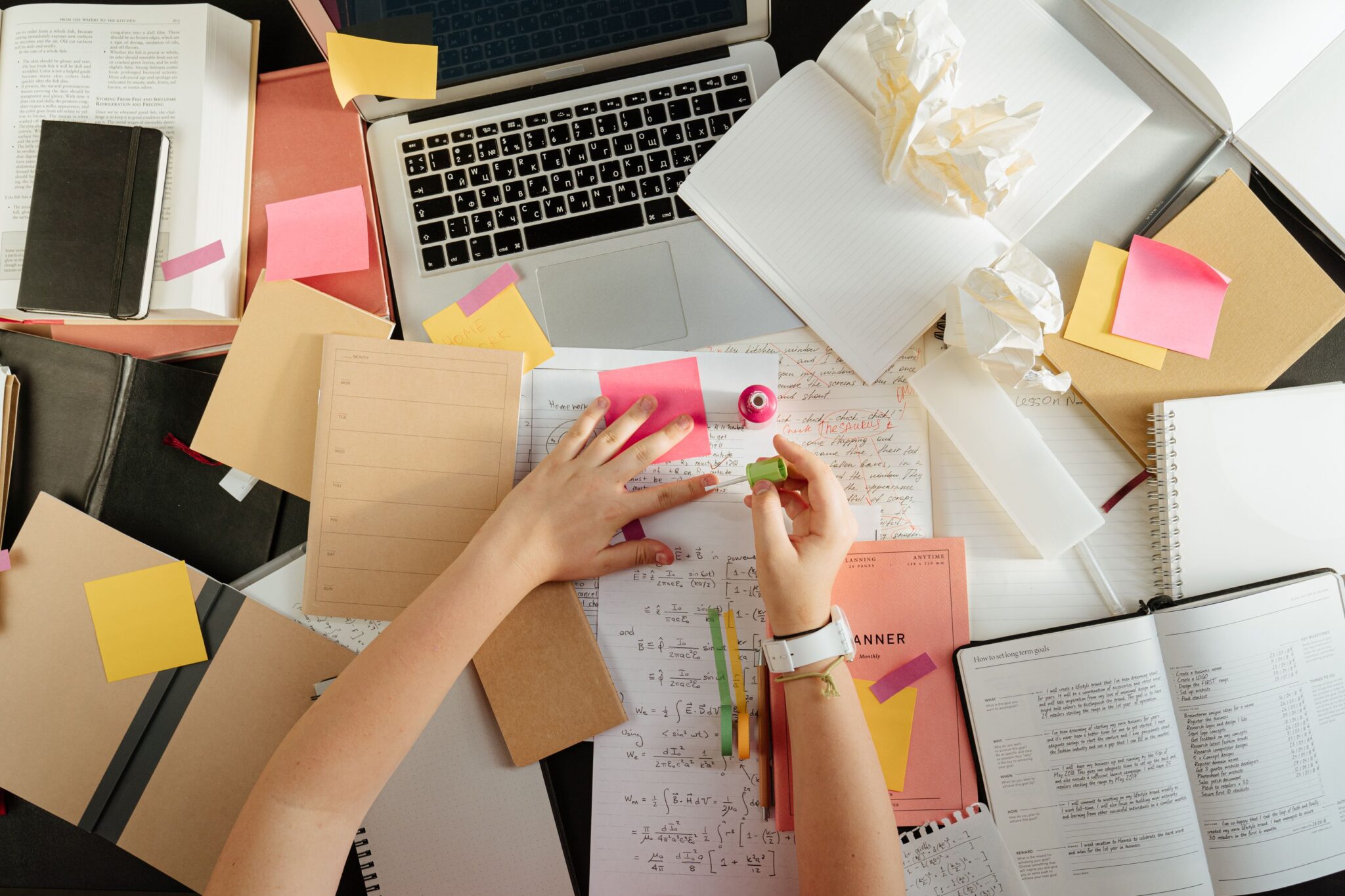 Overhead view of a cluttered desk with a laptop, notebooks, planners, books, sticky notes and handwritten papers scattered across the surface. A person is writing notes, with several crumpled sheets of paper nearby, suggesting stress, heavy workload or difficulty concentrating.