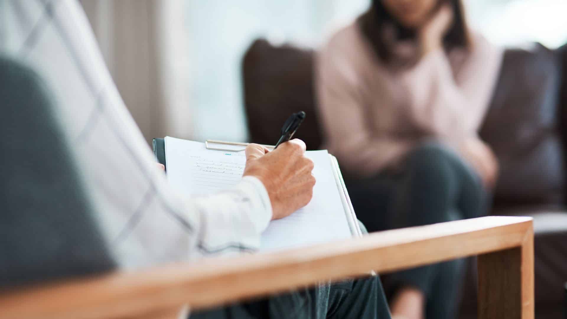 A person taking notes on a clipboard while talking with someone seated opposite them.