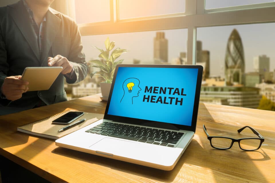 Image of a laptop on a desk with London in the background. The screen says mental health.
