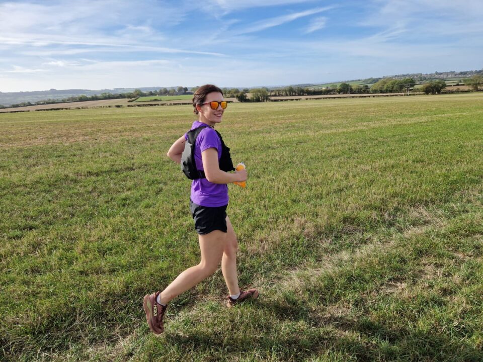 A woman in a purple running top and sunglasses runs across a grassy field, smiling towards the camera, with rolling countryside in the background.