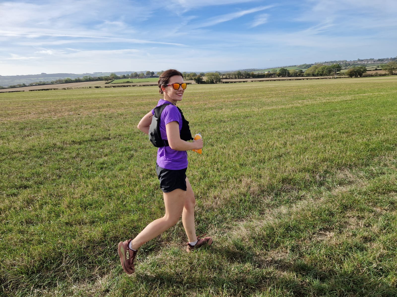 A woman in a purple running top and sunglasses runs across a grassy field, smiling towards the camera, with rolling countryside in the background.